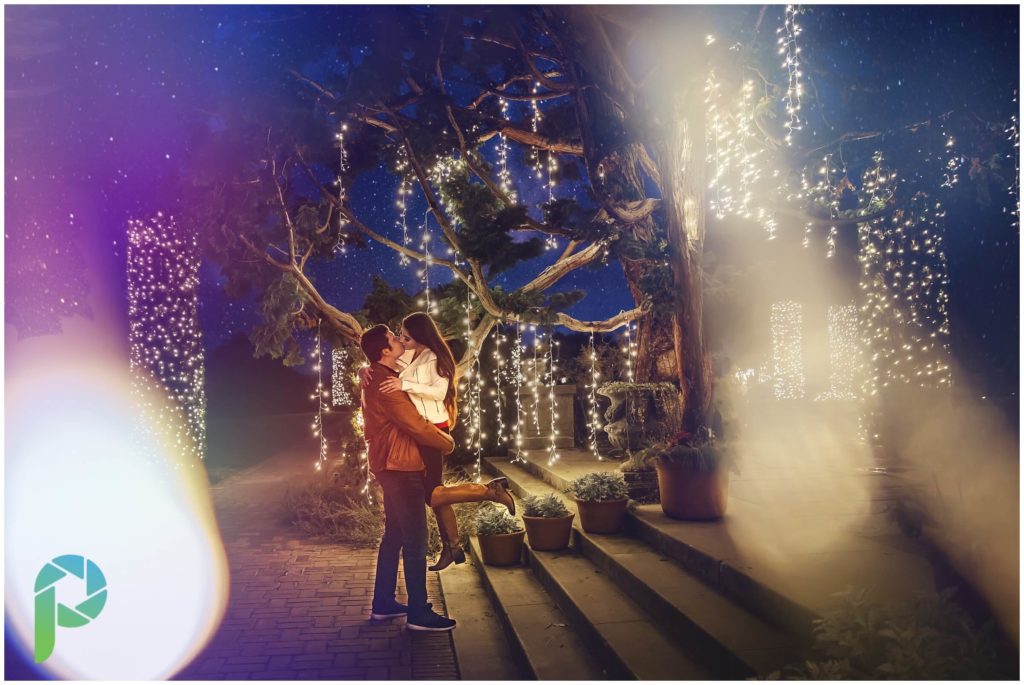 Man lifting up girl for a kiss in front of steps and christmas lights at Filoli in the San Francisco Bay Area at their holiday display.