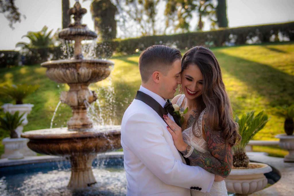 Bride and groom taking formal portraits at a Grand Island Mansion wedding in Walnut Grove, California. There is a fountain with running water in the background. The groom has his forehead to the bride's temple and the bride is looking down smiling.