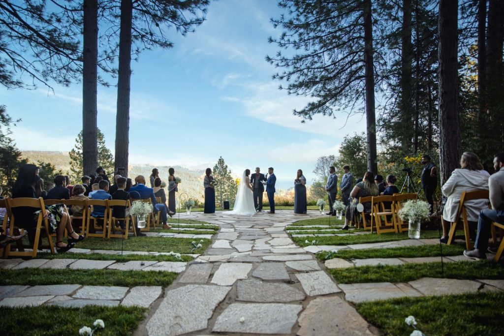 Photo taken at Forest House Lodge by Sacramento wedding photographer Philippe Studio Pro. Bride and groom stand with an officiant and wedding guests looking on at the end of an aisle.  They have blue skies above, large trees on either side of them, and a pebble aisle surrounded by green grass in front of them. The end of the aisle looks out onto the California mountains.