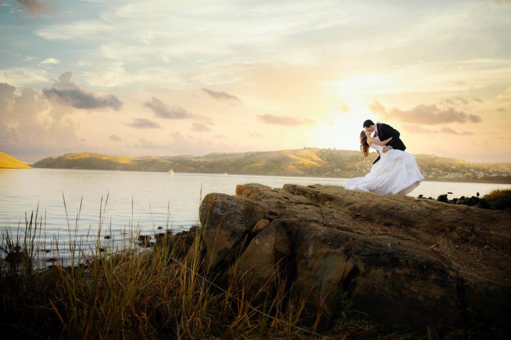 Image of groom dipping bride and tilting in for a kiss on a ledge by water's edge. To get good quality wedding photos such as this, the cost of photography for wedding may be more.