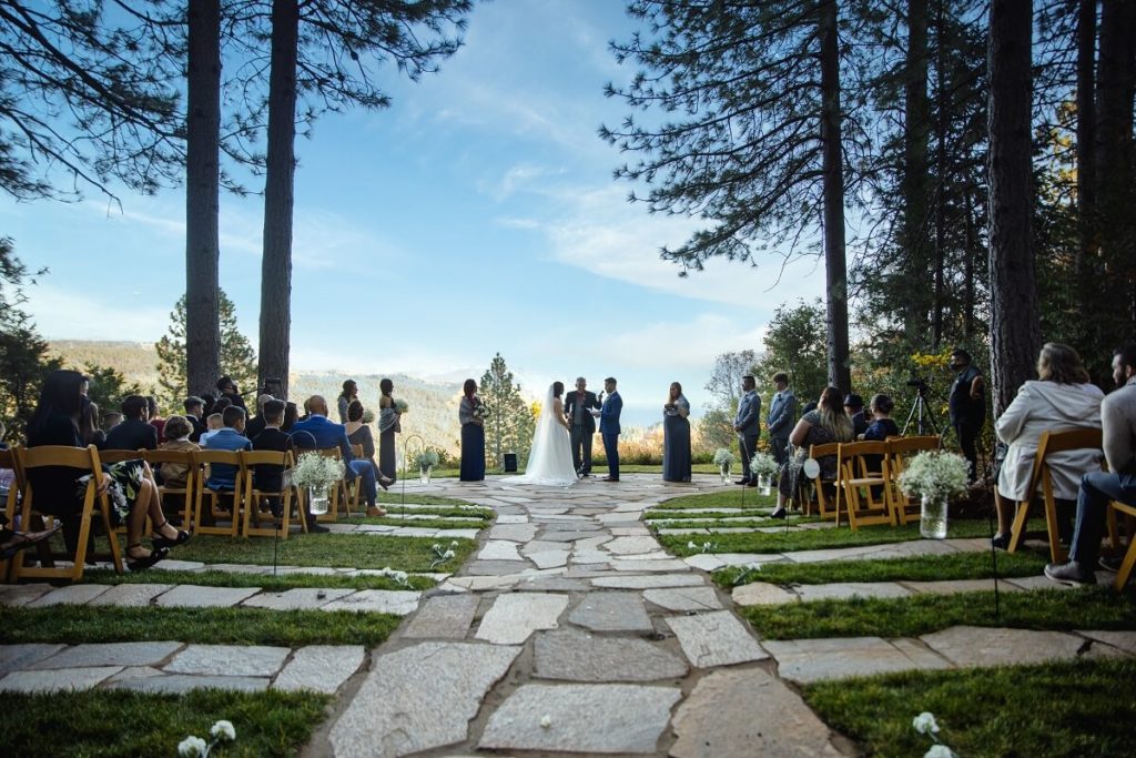 Wedding ceremony happening at forest house lodge with blue skies and the sierra mountains in the background. Bride and groom stand at the end of the alter with an officiant and guests looking on. One of the best outdoor wedding venues in Sacramento.
