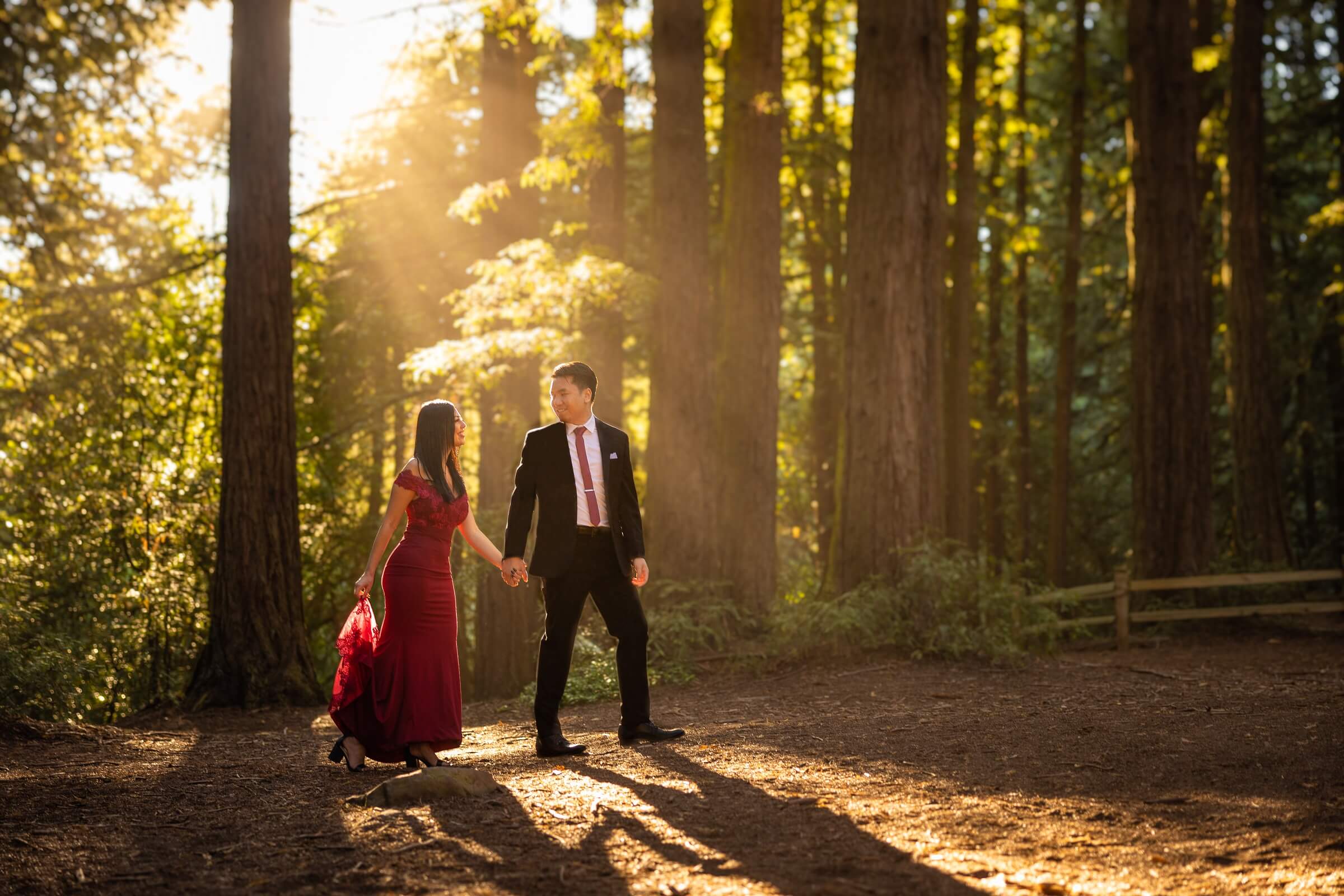 Engaged couple walk in wooded area of Reinhardt Redwood Forest. Groom looks back and bride, with sun shining through. photo by bay area engagement photographer philippe studio pro